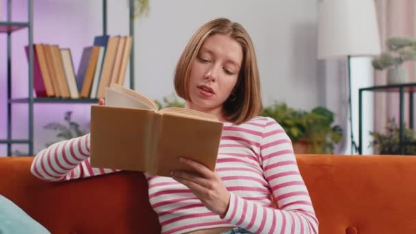 Młoda brunetka relaksująca interesującą książkę przewracając kartki uśmiechając się ciesząc literaturą odpoczywając na wygodnej sofie. Spokojny uśmiech wesoły dziewczyna w domu mieszkanie pokój dzienny siedzi na kanapie - Materiał filmowy, wideo