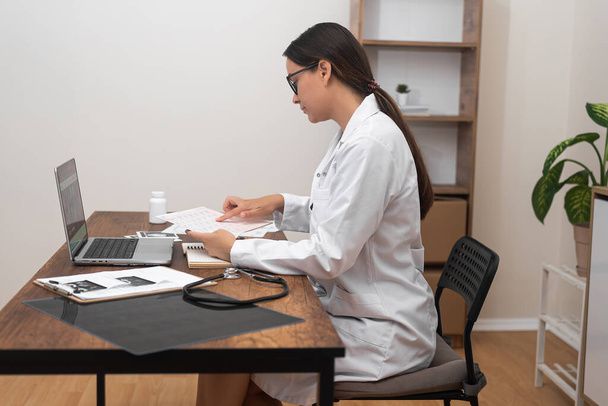 臨床記録解析:白いラボコートでは,女性医師が現代の診療所のテーブルで患者の医療文書を注意深く評価する.  - 写真・画像