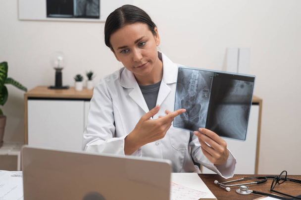 Молодий лікар розмовляє з пацієнтом онлайн через ноутбук, сидячи в офісі жінка вказує палець на зображення рентгенівського обстеження, пояснюючи особливості дистанційного лікування хвороби - Фото, зображення