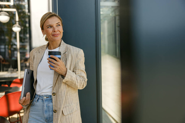 Femme d'affaires souriante buvant du café pendant la pause au bureau et regarde ailleurs  - Photo, image