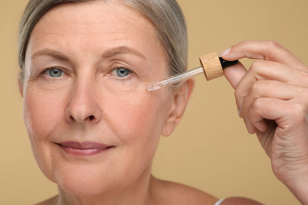Senior woman applying cosmetic product on her aging skin against beige background. Rejuvenation treatment - Foto, Bild