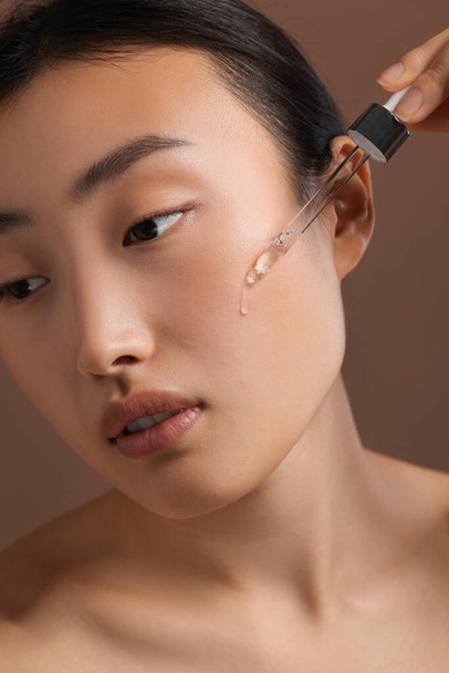 Beautiful young woman applying cosmetic serum onto her face on brown background, closeup - 写真・画像