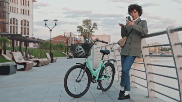 Σύγχρονη επιχειρηματίας με ποδήλατο στέκεται χαλαρή σε εξωτερικούς χώρους το πρωί μιλάμε για βιντεοκλήση στο smartphone - Πλάνα, βίντεο