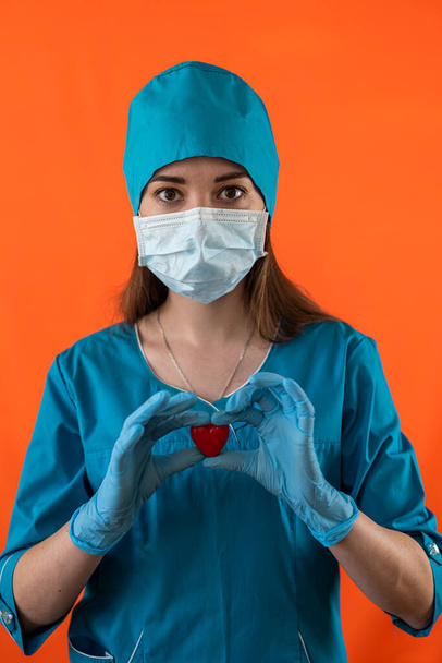 Nuori naislääkäri sairaalan vaatteissa naamio ja käsineet pitämällä pieni punainen sydän symbolina sairaalassa. Operatiivinen. Hoitaja. Sydänsymboli - Valokuva, kuva