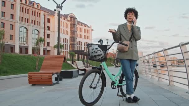 Длинный снимок современной деловой женщины в стильной умной повседневной одежде, стоящей с велосипедом на открытом воздухе, разговаривающей по телефону рано утром - Кадры, видео
