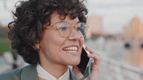 Zbliżenie portret radosnej białej kobiety z kręconymi włosami noszącej okulary stojące na zewnątrz po rozmowie telefonicznej - Materiał filmowy, wideo