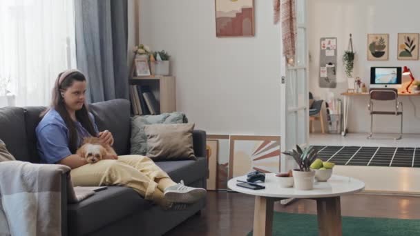 Современная молодая женщина с синдромом Дауна сидит на диване в гостиной дома, гладит собаку и разговаривает с кем-то по телефону - Кадры, видео