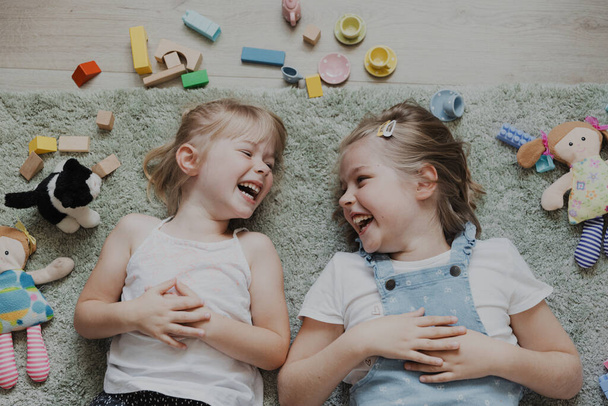 Κάτοψη από ψηλά για τα παιδιά που παίζουν με πολύχρωμα παιχνίδια. Πορτρέτο των παιδιών που βρίσκονται στο χαλί στο σπίτι υπνοδωμάτιο ή νηπιαγωγείο. Δύο χαρούμενες χαμογελαστές αδερφές. Εκπαιδευτικά φιλικά προς το περιβάλλον παιχνίδια - Φωτογραφία, εικόνα