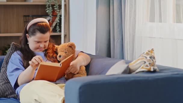 Schwenk-Aufnahme eines jungen Mädchens mit Down-Syndrom, das sich auf dem Sofa entspannt, Spielzeugbär umarmt und Roman liest - Filmmaterial, Video