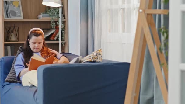 Panning záběr moderní mladé bělošky s Downovým syndromem relaxační na gauči čtení knihy - Záběry, video