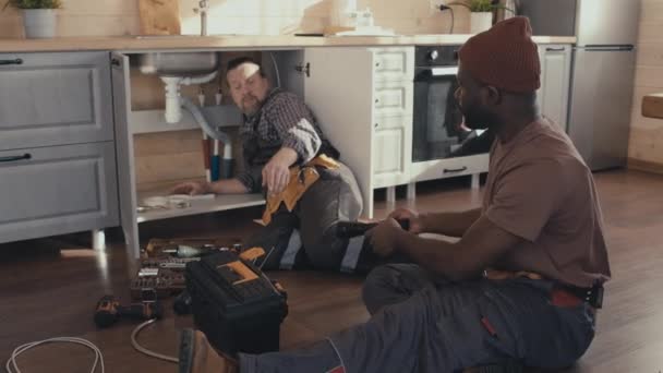 キッチンに床に座っている若いアフリカ系アメリカ人の修理士は,彼の成熟したコーカサス人の同僚にシンクの排水を修理するツールを与えます - 映像、動画