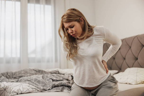 Беременная женщина дышит и держит спину от боли, сидя на кровати в спальне.  - Фото, изображение