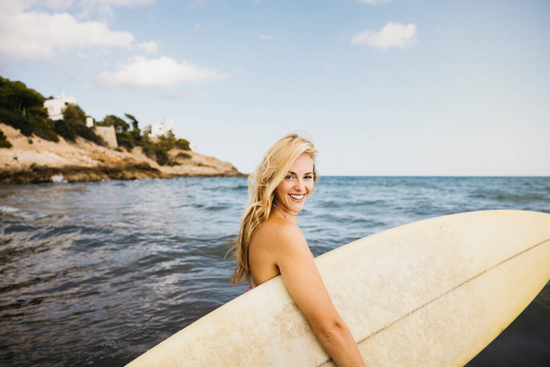 Mooie glimlachende blonde vrouwen in zwemkleding met een surfplank aan het strand tijdens een leuke vakantie aan zee. - Foto, afbeelding