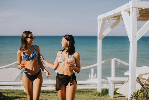 Две улыбающиеся девушки в бикини наслаждаются отдыхом на пляже, выпивая коктейль - Фото, изображение