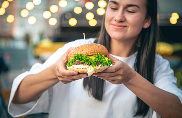 Μια ελκυστική νεαρή γυναίκα στα χέρια κατέχει ένα burger, τρώει fast food στο δρόμο καφέ σε ένα θολό φόντο. - Φωτογραφία, εικόνα