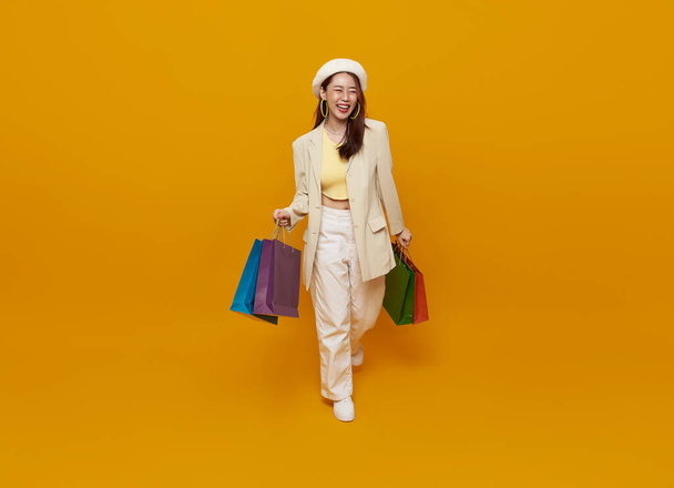 Happy Asian woman carrying colorful shopping bags in shopping mall center isolated on yellow studio background. - Fotó, kép