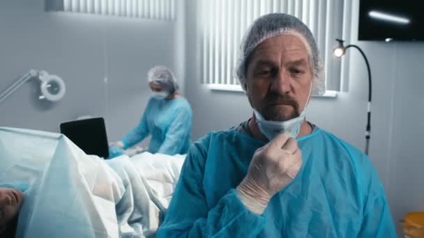 Retrato de enfoque selectivo del cirujano varón maduro de pie en el quirófano quitándose la máscara después de terminar la operación - Metraje, vídeo