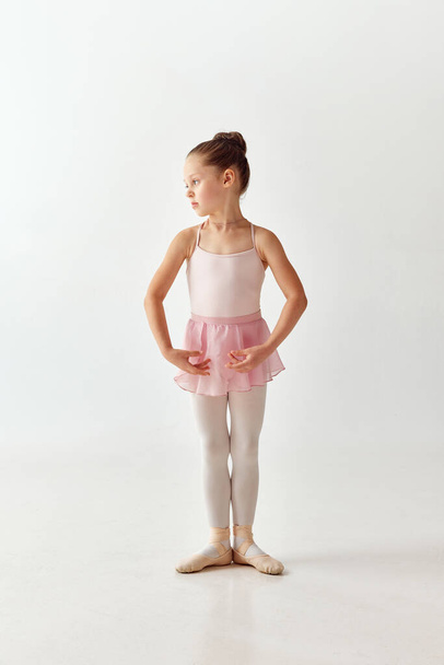 Портрет маленькой очаровательной балерины-танцовщицы в балетном платье из розовой пачки и в пуантах с белыми ногами, стоящей на белом фоне. Концепция красоты, моды, хобби, самовыражения - Фото, изображение