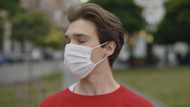Serieuze mannelijke portret dragen medische witte masker buiten met City Achtergrond. Blanke man, jongen, student, toerist staat en kijkt naar de camera tijdens de pandemie. Sluiten - Video