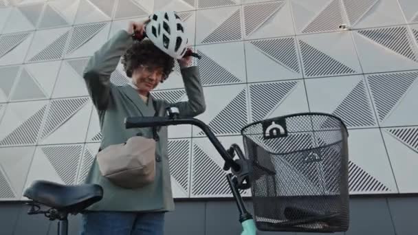 Low angle view shot of Caucasian woman with curly hair wearing smart casual clothes putting on helmet then going by bicycle - Footage, Video