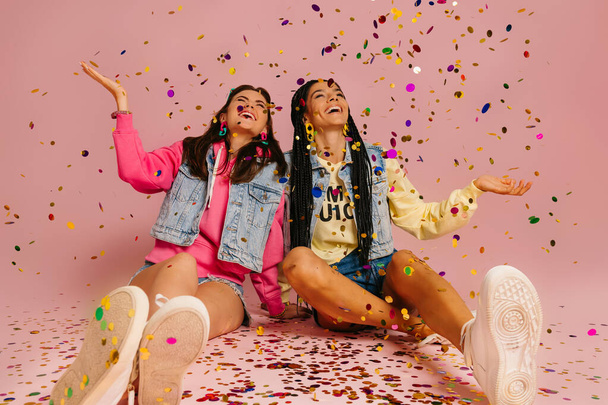 Two joyful young women throwing colorful confetti while sitting against pink background - Foto, imagen