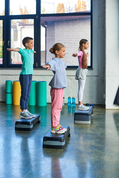 three adolescent girls and boy in sportswear exercising on fitness stepper with arms asides, sport - Photo, Image