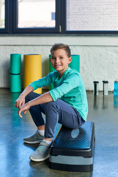 radosny chłopiec w odzieży sportowej siedzi na kroku fitness i uśmiecha się szczęśliwie do kamery, sport dla dzieci - Zdjęcie, obraz