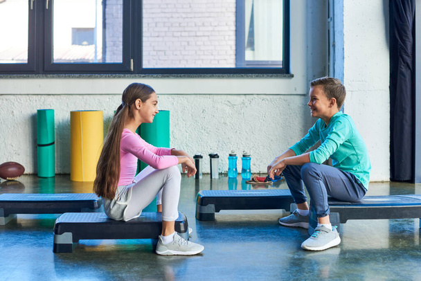 wesoły chłopiec i dziewczyna siedzi na fitness steppers i uśmiechając się szczęśliwie do siebie, sport dziecko - Zdjęcie, obraz