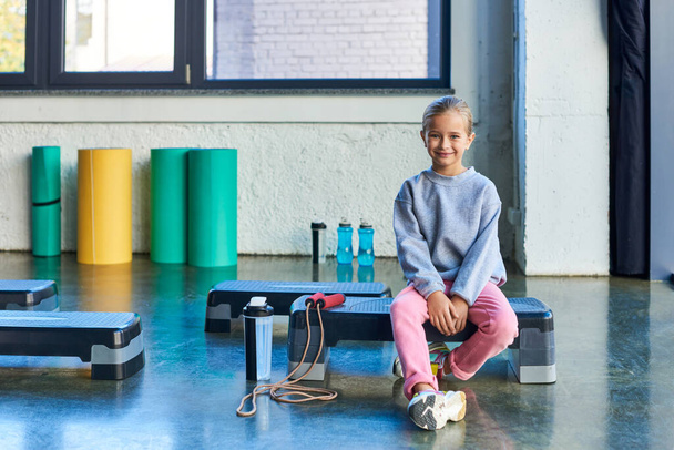 alegre rubia niña sentada en fitness stepper en ropa deportiva sonriendo a la cámara, deporte infantil - Foto, imagen