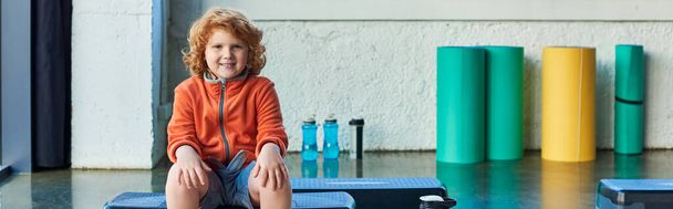 niño de pelo rojo feliz sentado en el paso de fitness sonriendo alegremente a la cámara, deporte infantil, pancarta - Foto, imagen