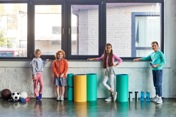 quatre garçons et filles mignonnes pré-adolescentes en vêtements de sport posant par la fenêtre dans la salle de gym, sport pour enfants - Photo, image