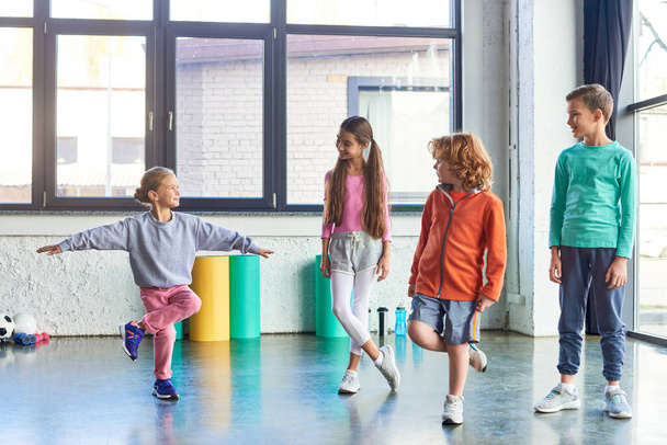 μικρό ξανθό κορίτσι στέκεται στο ένα πόδι τεντώνεται μπροστά από άλλα χαρούμενα παιδιά, παιδικό άθλημα - Φωτογραφία, εικόνα