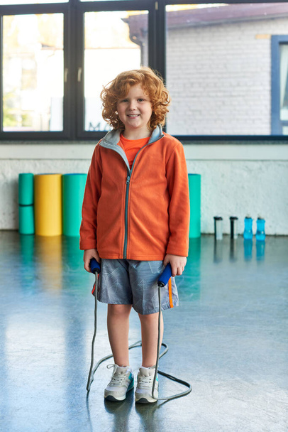iloinen punatukkainen poika poseeraa hyppyköyden kanssa ja hymyilee iloisesti kameralle, lapsiurheilu - Valokuva, kuva