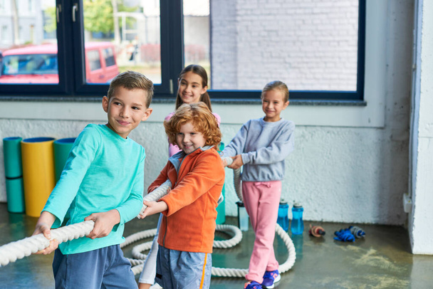 веселые красивые дети в спортивной одежде тянут канат в спортзале и весело улыбаются, детский спорт - Фото, изображение