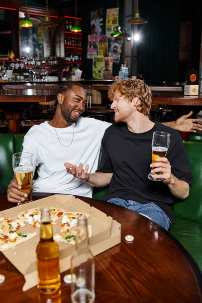 счастливый рыжий мужчина жестикулирует в чате с африканским американским другом за пивом и пиццей в баре - Фото, изображение