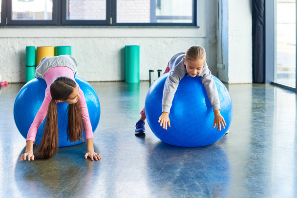 two joyful girls in sportswear lying on fitness balls in gym stretching hands to floor, child sport - Photo, Image