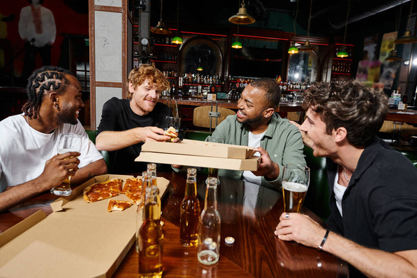 четыре счастливых многонациональных друга делятся пиццей и пьют пиво в баре, мужчины на мальчишнике - Фото, изображение