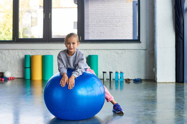 красивая маленькая девочка в спортивной одежде лежит на фитнес-мяч и слегка улыбается на камеру, детский спорт - Фото, изображение