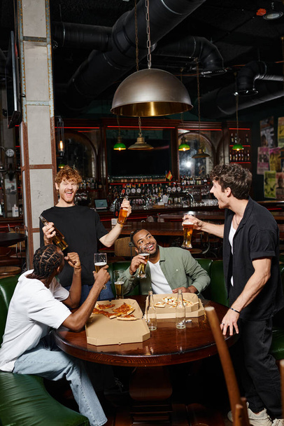 возбужденные межрасовые друзья тосты с бутылками и стаканами пива в баре, проводить время вместе - Фото, изображение