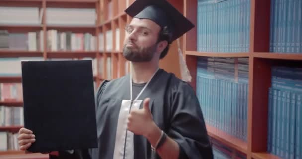 幸せな男性学生は,彼の卒業証書を保持しながら,それを達成するために幸せな親指を示しています. 大学図書館で彼の業績を披露する誇りある学生 - 映像、動画