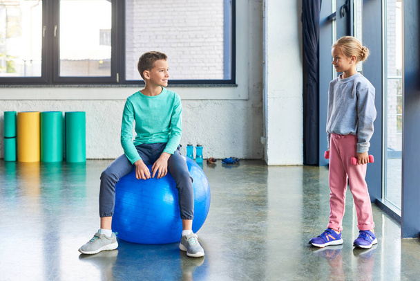 little blonde girl with dumbbells in hands smiling at cute boy on fitness ball, child sport - Photo, Image