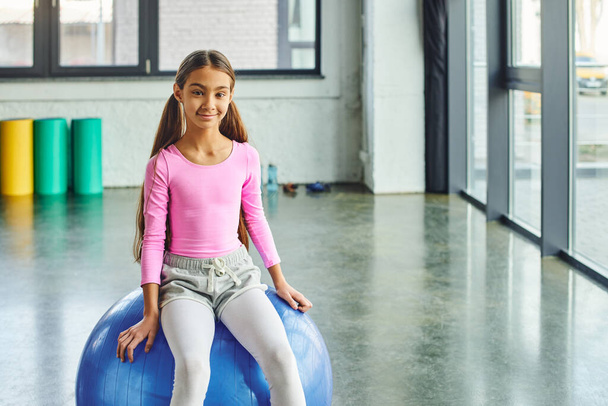 веселая милая девушка с длинными волосами сидя на фитнес-мяч и глядя на камеру, детский спорт - Фото, изображение