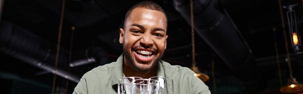 sztandar szczęśliwy Afroamerykanin człowiek z aparatem na zębach trzyma trzy szklanki w barze, zabawy - Zdjęcie, obraz