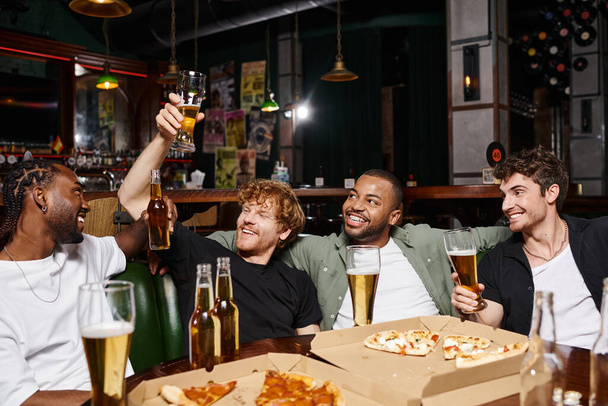 four happy interracial friends raising bottles and glasses of beer in bar, spending time together - Photo, Image