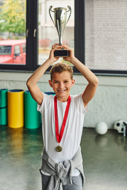 vertical shot of cheerful little boy with golden medal raising trophy above his head, child sport - Photo, Image