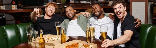 pancarta de alegres amigos interracial abrazando y sentado cerca de pizza y cerveza, pasar tiempo en el bar - Foto, Imagen
