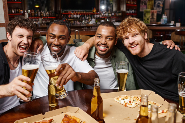 grupa szczęśliwych wielonarodowych przyjaciół przytulających się i wznoszących toast szklankami piwa, spędzających czas w barze - Zdjęcie, obraz