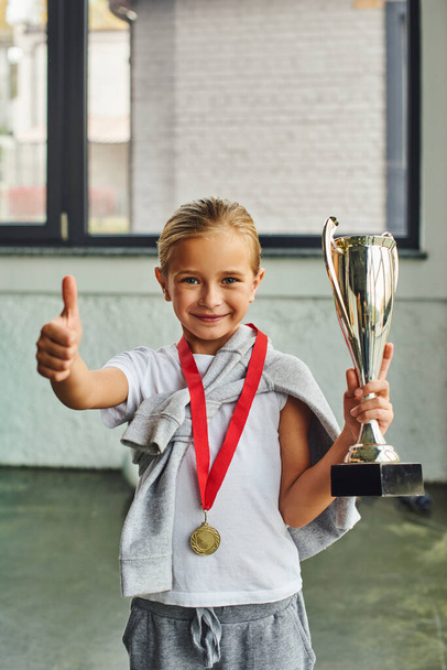 tiro vertical de chica rubia con trofeo y medalla de oro que muestra el pulgar hacia arriba en la cámara, deporte infantil - Foto, imagen