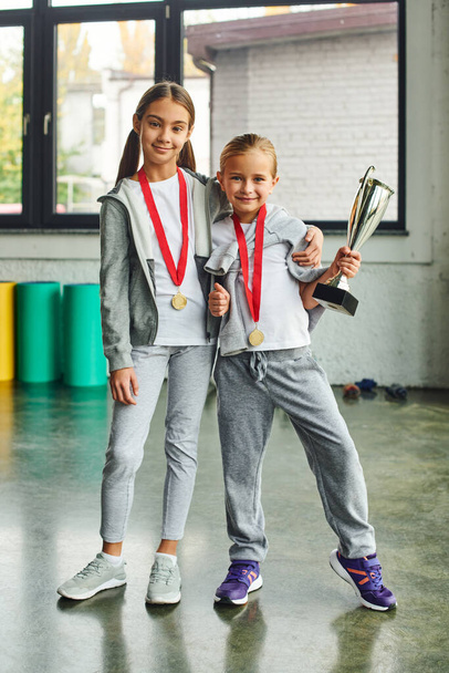 vertical shot of cute preadolescent girls with medals and trophy hugging and smiling at camera - Photo, Image