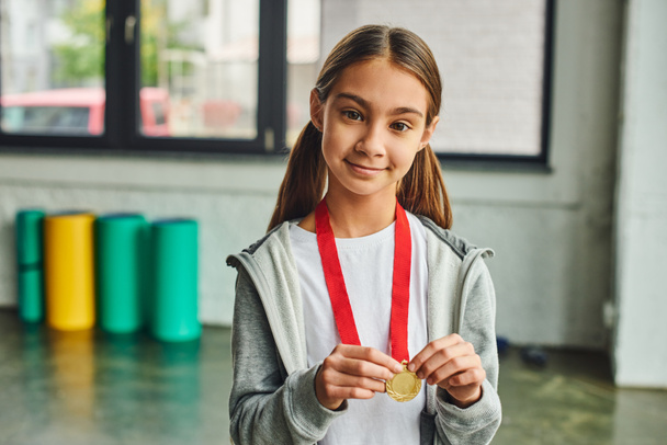 Smiley hübsches Mädchen mit langen Haaren hält goldene Medaille in der Hand und schaut freudig in die Kamera, Kindersport - Foto, Bild